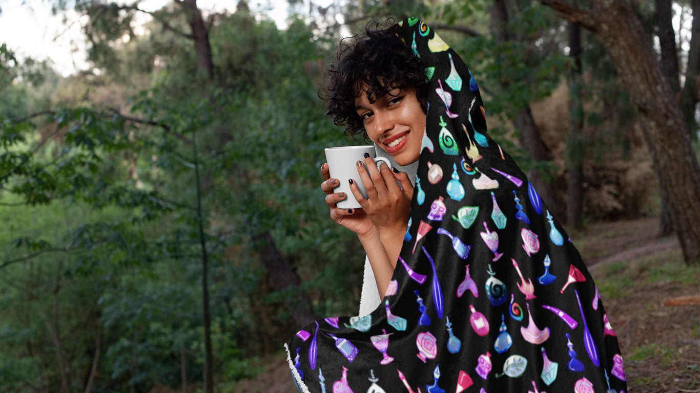 A young woman with a coffee cup sitting in the forest, wearing a hooded blanket, covered in multicolored potion bottles